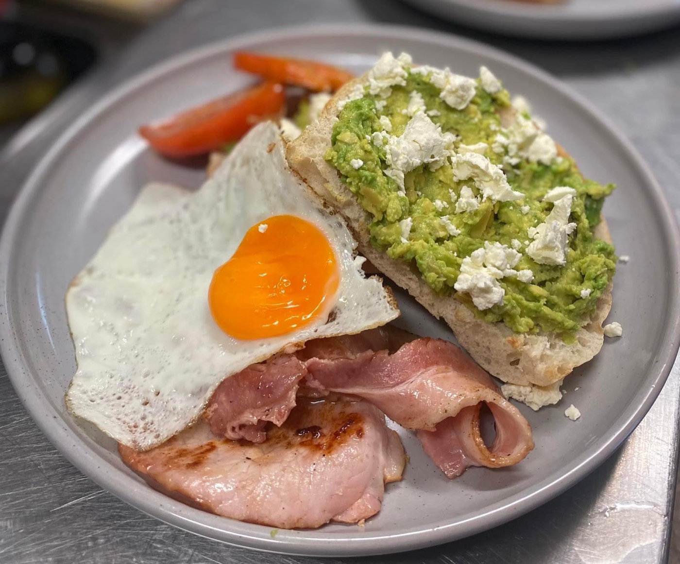 Smashed avocado with crumbled feta on toast, a fried egg and rashers of bacon on a grey plate
