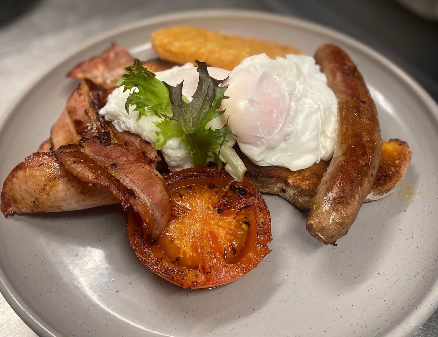 A cooked breakfast on a grey plate: a poached egg on salad leaves, a sausage, bacon, a grilled tomato and a hash brown