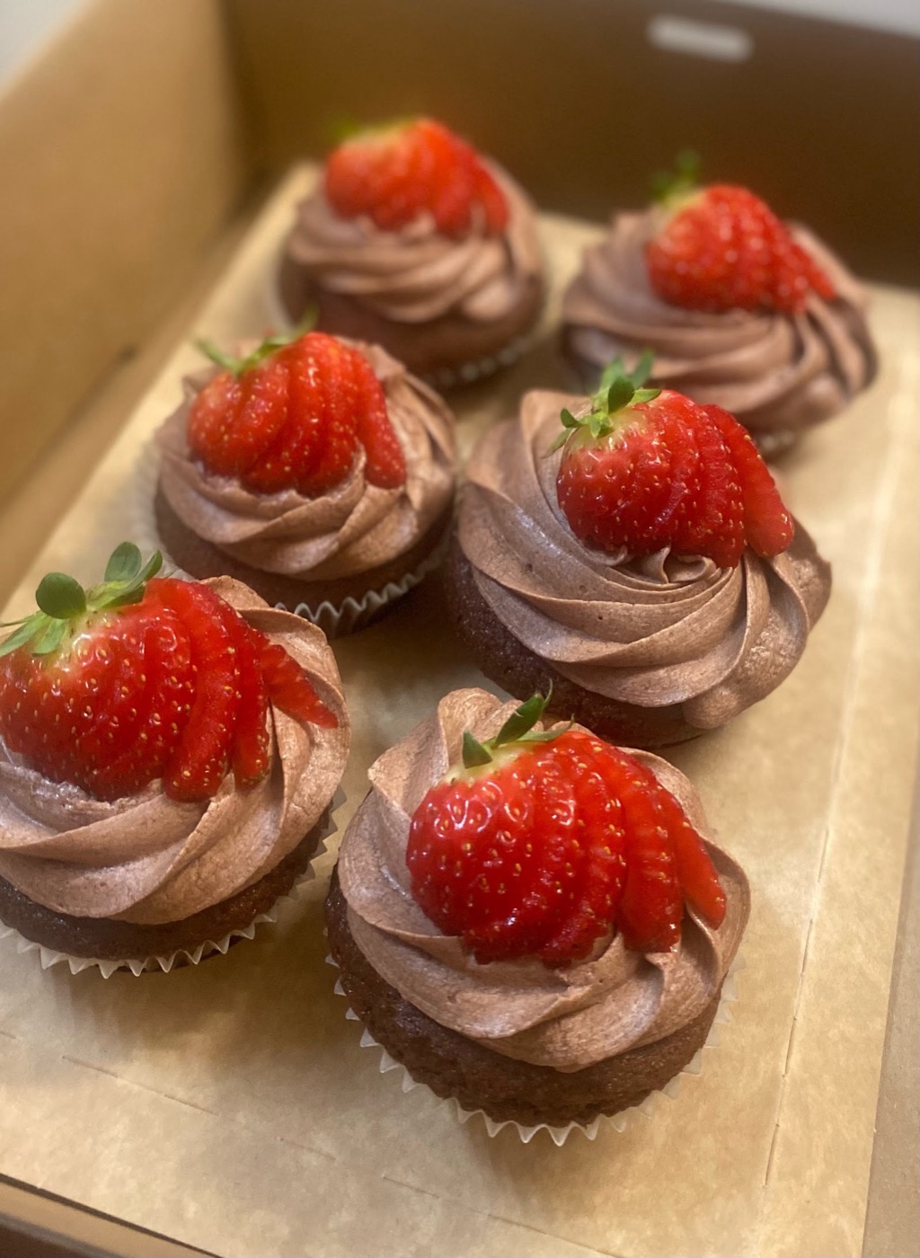A box of six chocolate cupcakes with pink icing, each topped with a fresh strawberry
