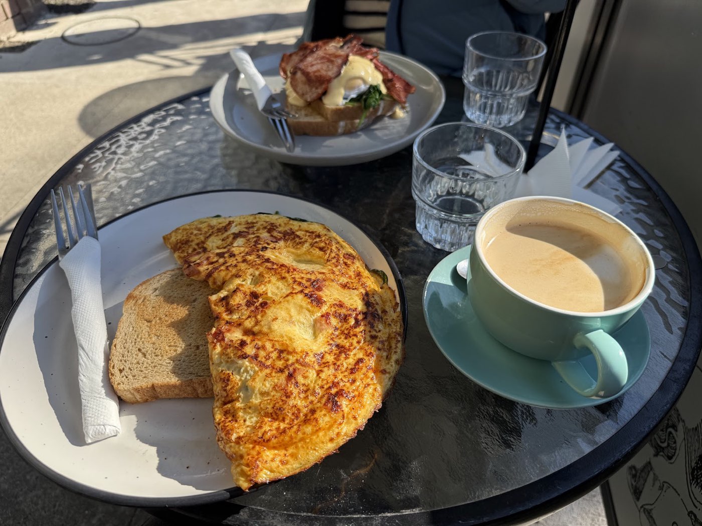 A folded omelette with toast and a cup of coffee on a glass table outside, with a plate of eggs benedict behind