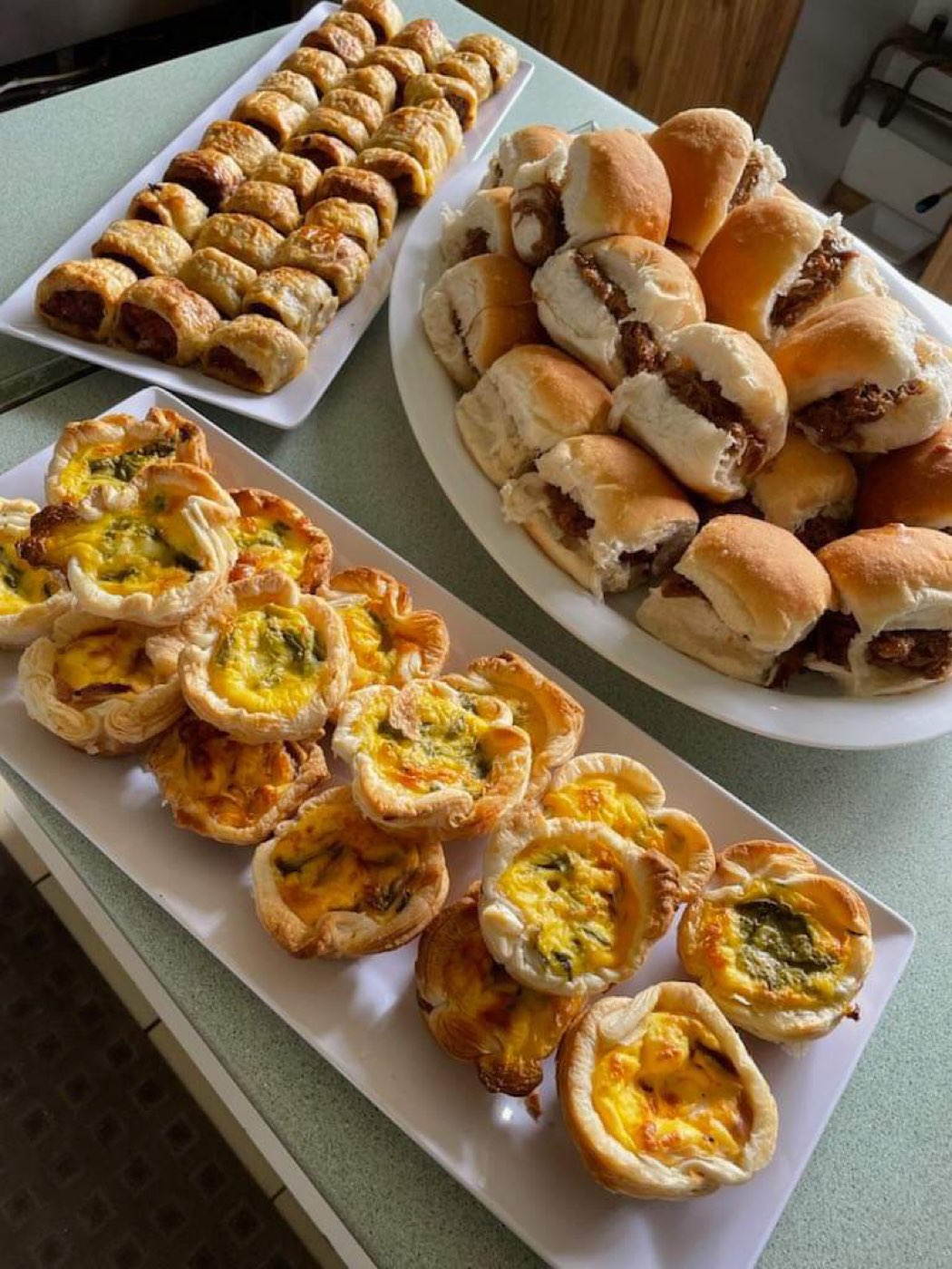 Platters of mini quiches, sausage rolls and small filled bread rolls on a long tray