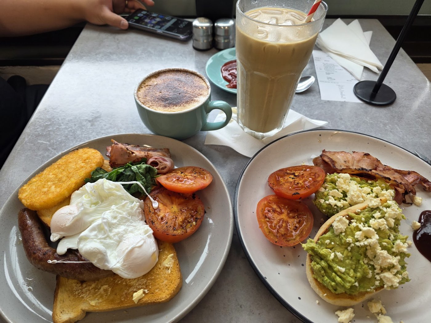 Two breakfast plates on a table beside a flat white and an iced coffee: one with poached eggs, hash brown and toast, one with smashed avocado, bacon and tomato